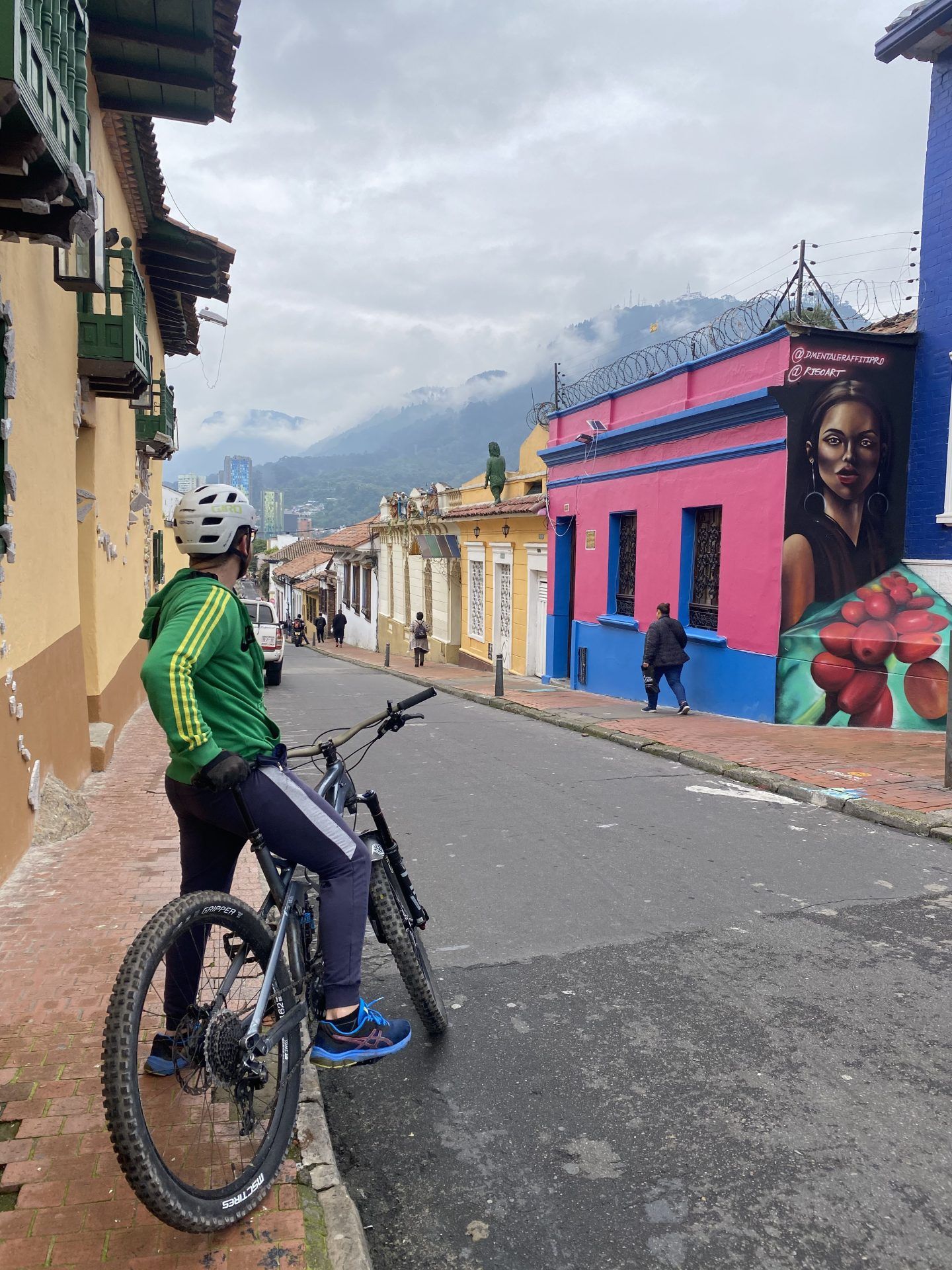 A cyclist in a white helmet and green jacket stops his mountain bike on a La Candelaria street lined with colorful houses and a large mural, with cloud-covered hills behind