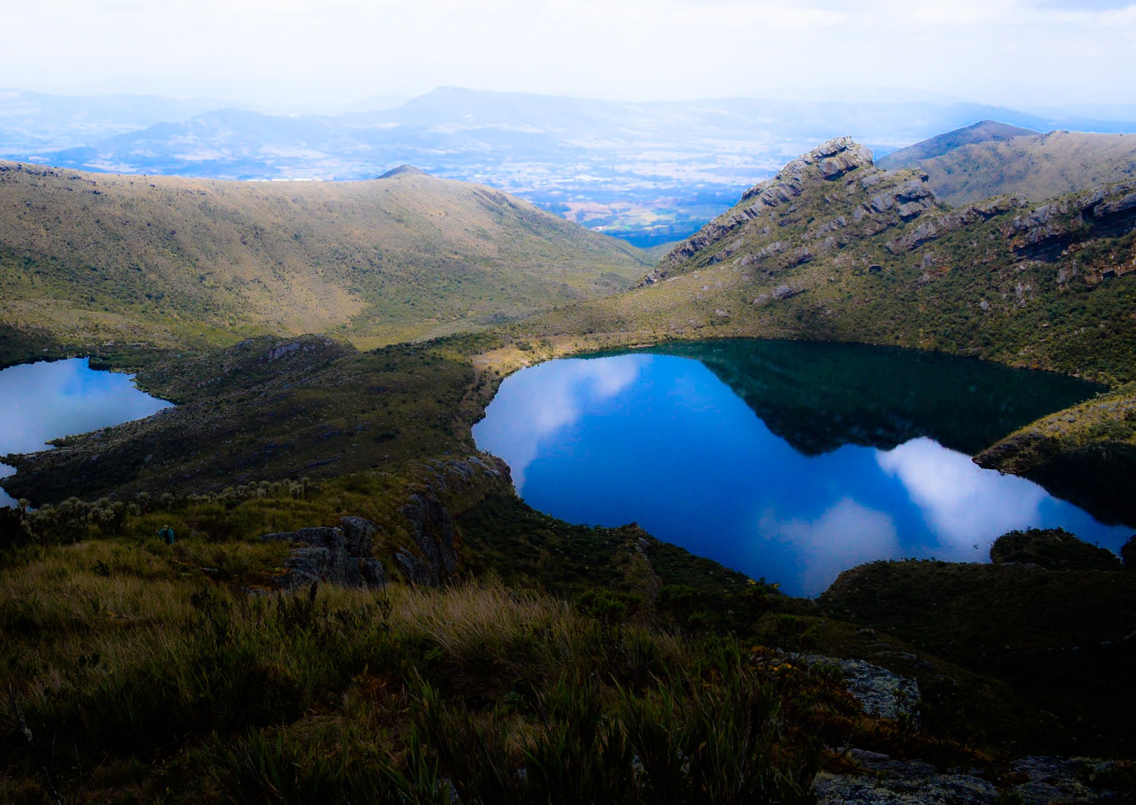 Una de las lagunas de Siecha, de agua azul oscura que refleja nubes, rodeada de laderas de páramo y peñascos, con otra laguna a la izquierda y el valle al fondo