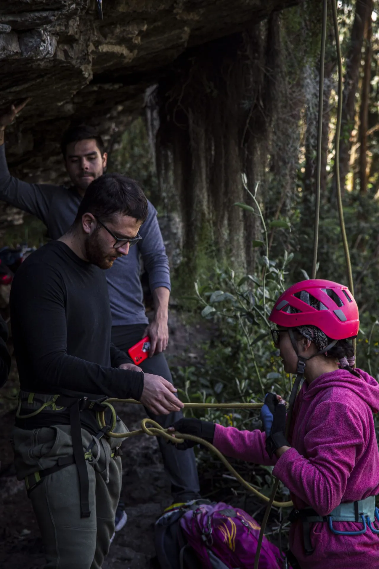 Una escaladora con casco rosado revisa el nudo de encordamiento en el arnés de un compañero bajo un techo de roca en Suesca