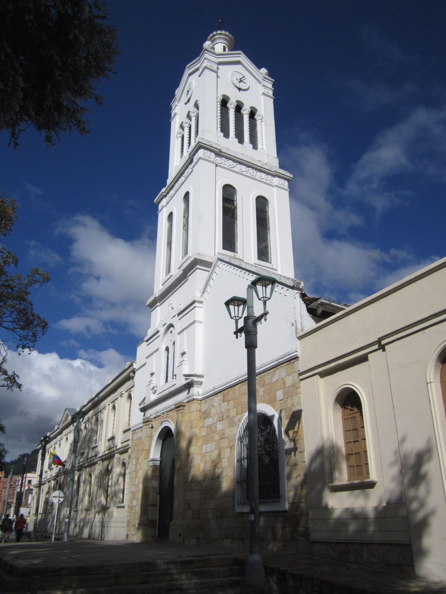 White bell tower of the Santa Bárbara church in Usaquén, with a clock near the top, a sandstone base and a deep blue sky behind it